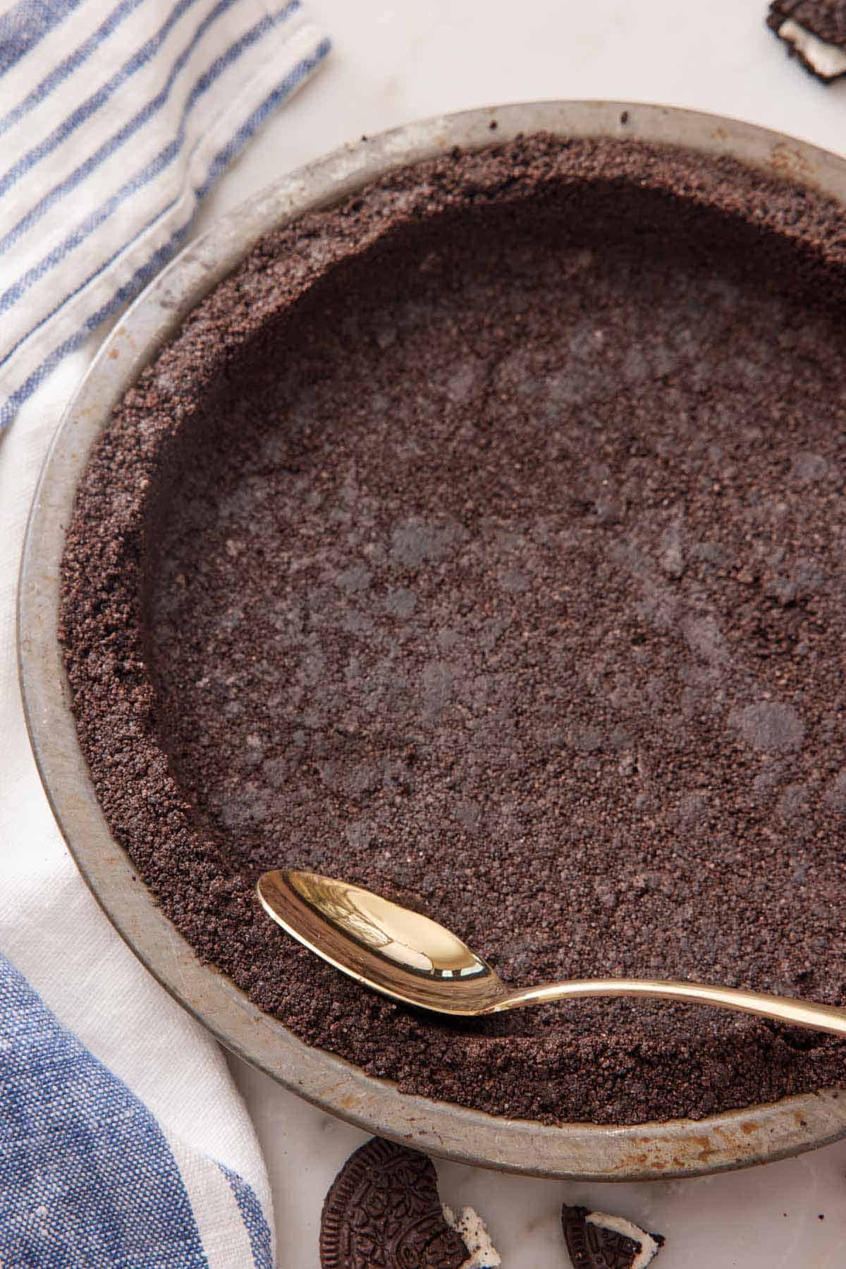 An Oreo pie crust in a pie pan with a spoon pressing the edge.