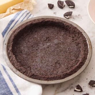 An Oreo pie crust in a baking pan with some topping, Oreos, and butter in the background.
