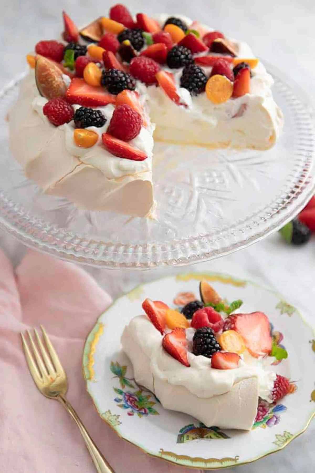 A plate with a slice of pavlova topped with berries with the rest of the pavlova on a clear cake stand.