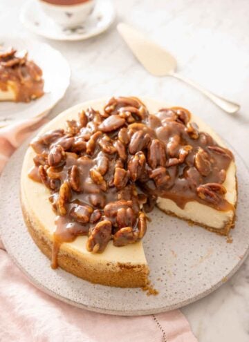 A pecan pie cheesecake with a slice taken out. A cake spatula and a cup of tea in the background.