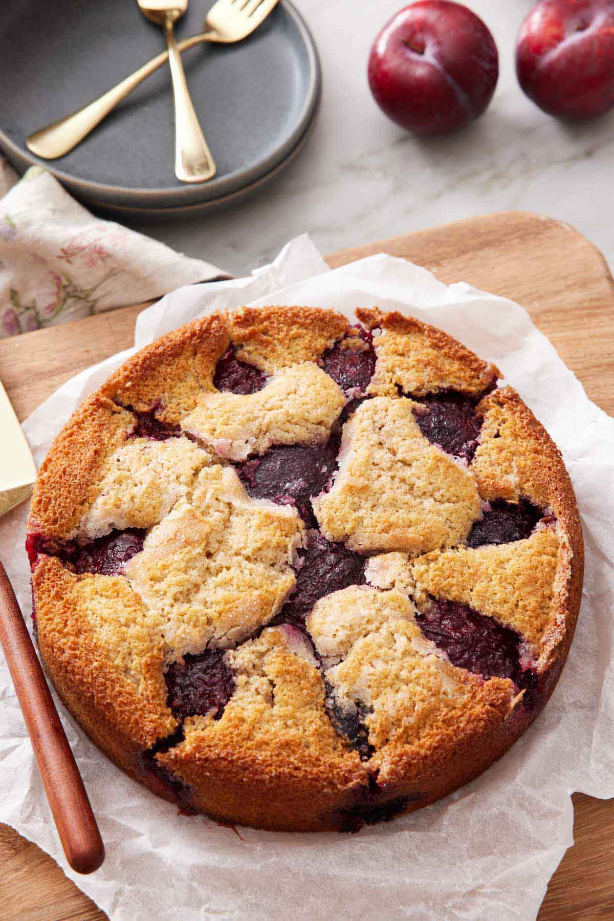 A plum cake on a parchment lined serving board. Some plums in the background with a stack of plates and forks.