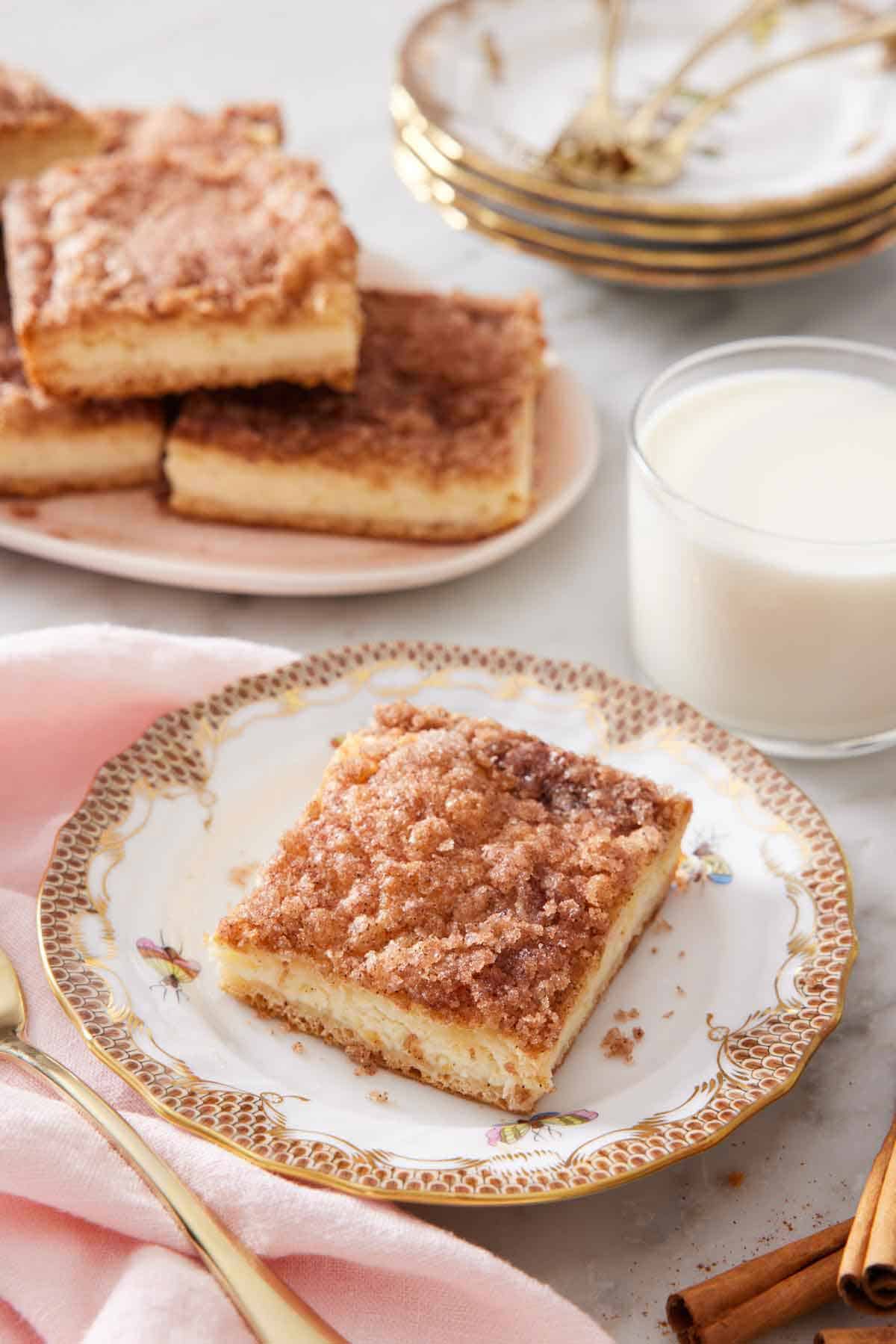 A square slice of a sopapilla cheesecake with a glass of milk and additional slices in the background.
