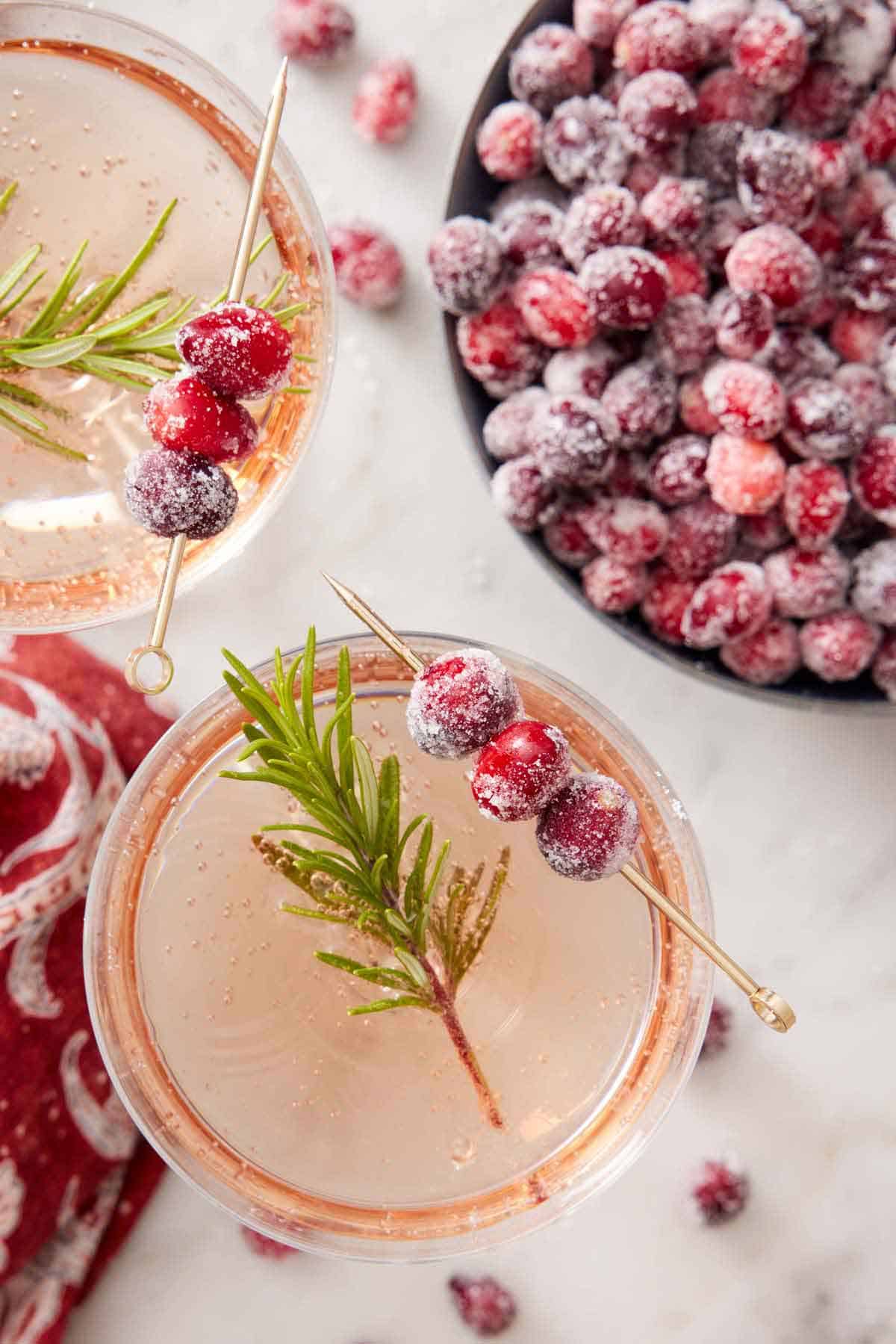 Overhead view of cocktails with a rosemary and sugared cranberries garnish. A bowl of more sugared cranberries on the side.
