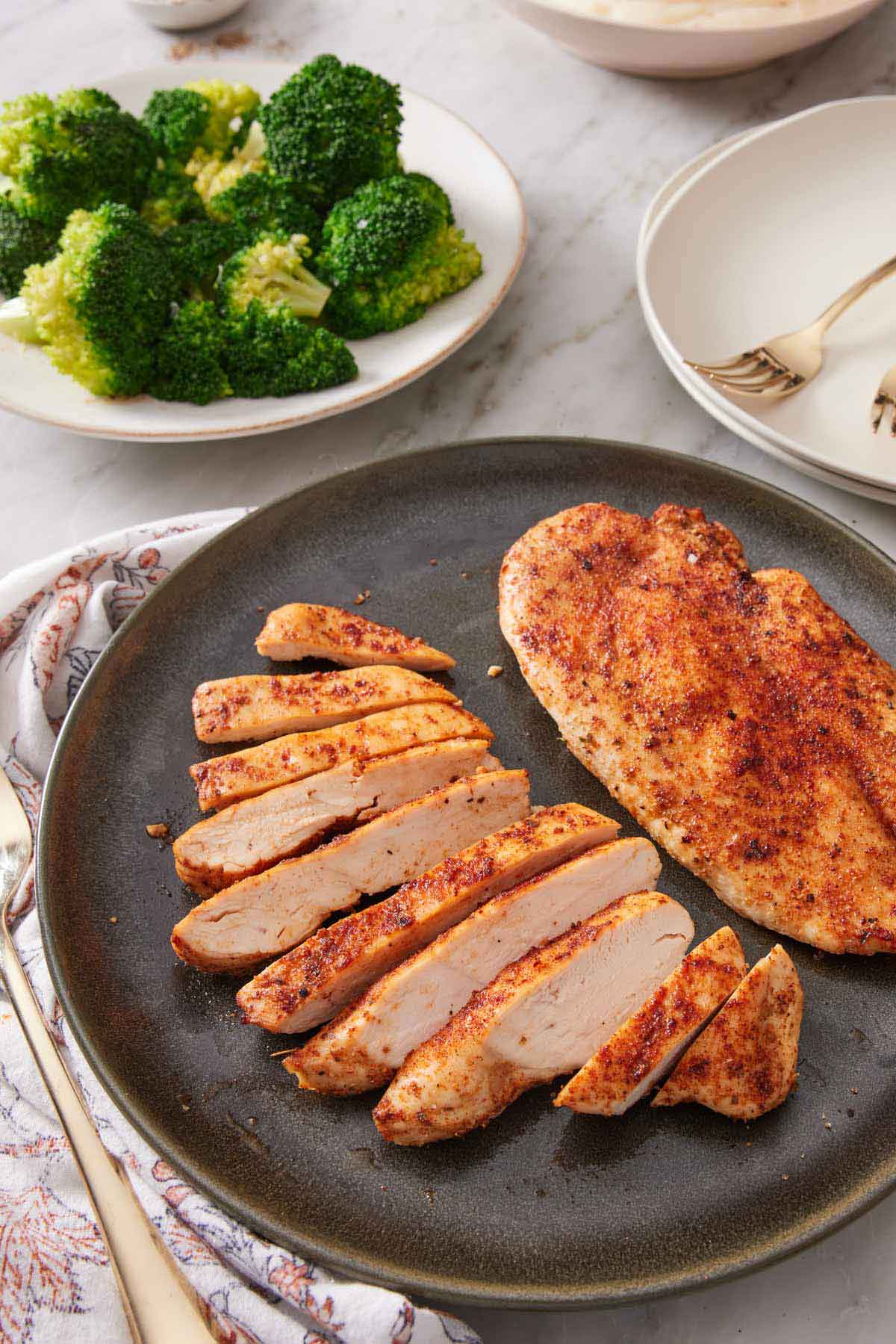 A plate with two air fryer chicken breasts, one breast sliced. Plate of broccoli in the background.