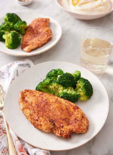 Two plates of air fryer chicken breasts with a side of broccoli. A drink and bowl of mashed potatoes in the background.