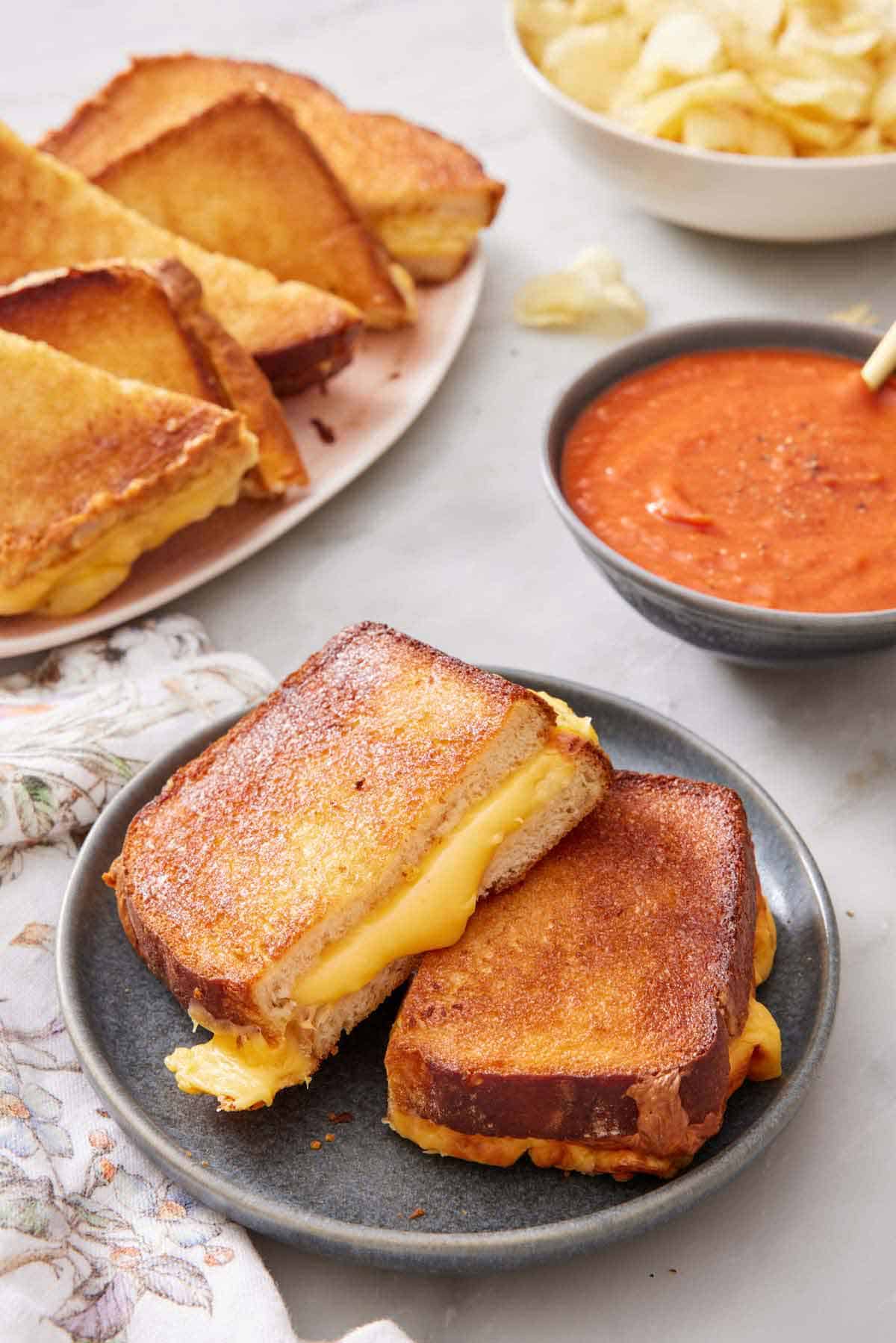 A plate with air fryer grilled cheese cut in half with more on a platter in the background along with a bowl of tomato soup.