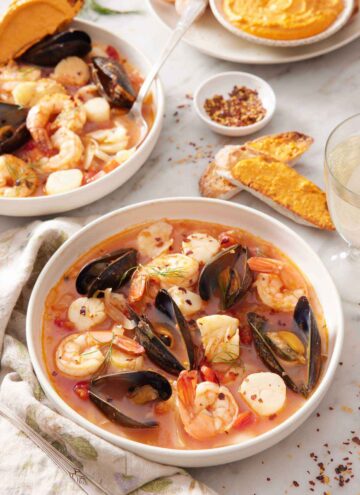 Two bowls of bouillabaisse, one in front, and bread with rouille spread on it in the background along with a bowl of rouille.