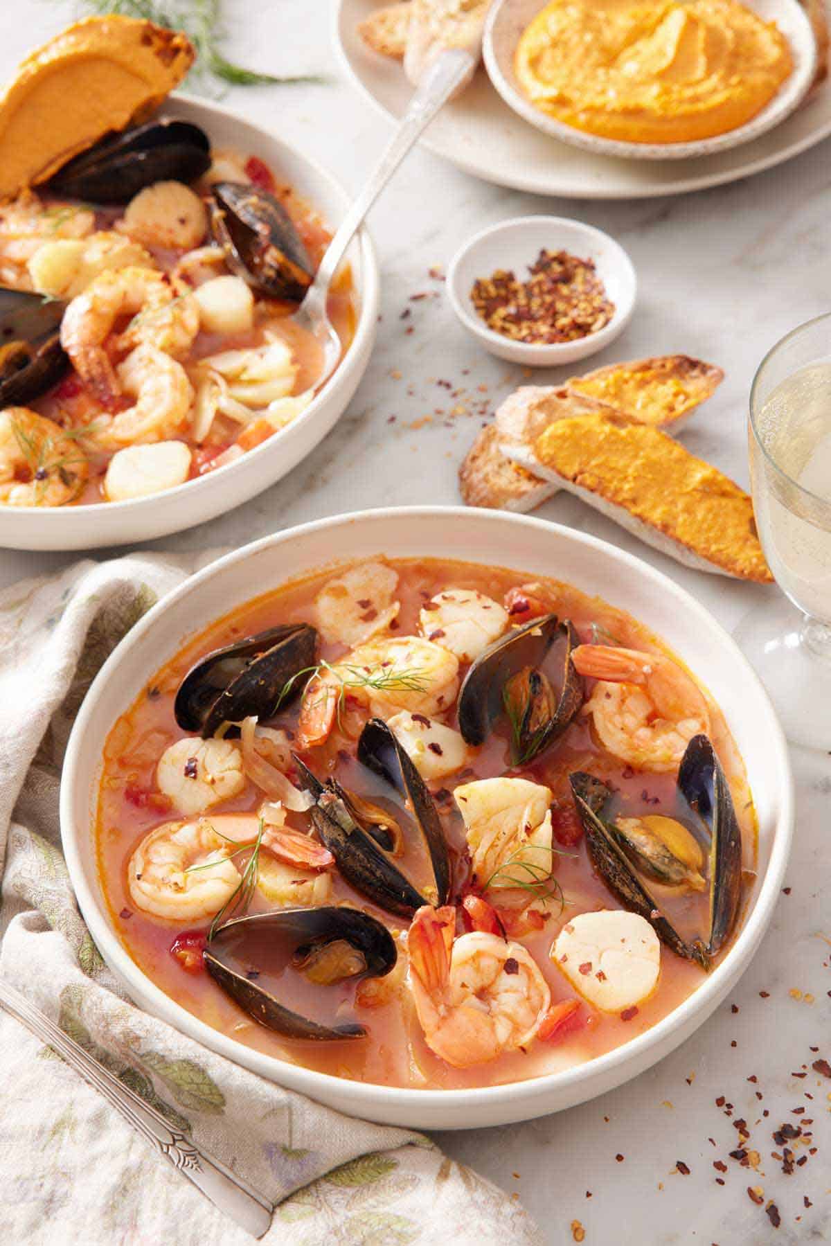 Two bowls of bouillabaisse, one in front, and bread with rouille spread on it in the background along with a bowl of rouille.