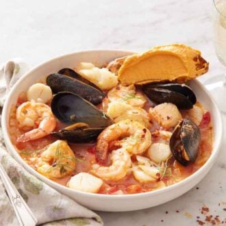 A bowl of bouillabaisse with a piece of bread with rouille on it and a glass of wine in the background.