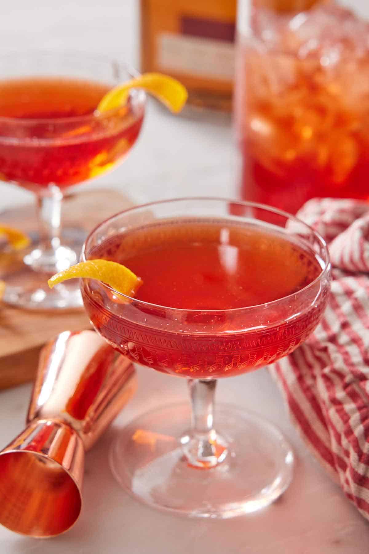 A glass of boulevardier with a jigger beside it. A second cocktail in the background.