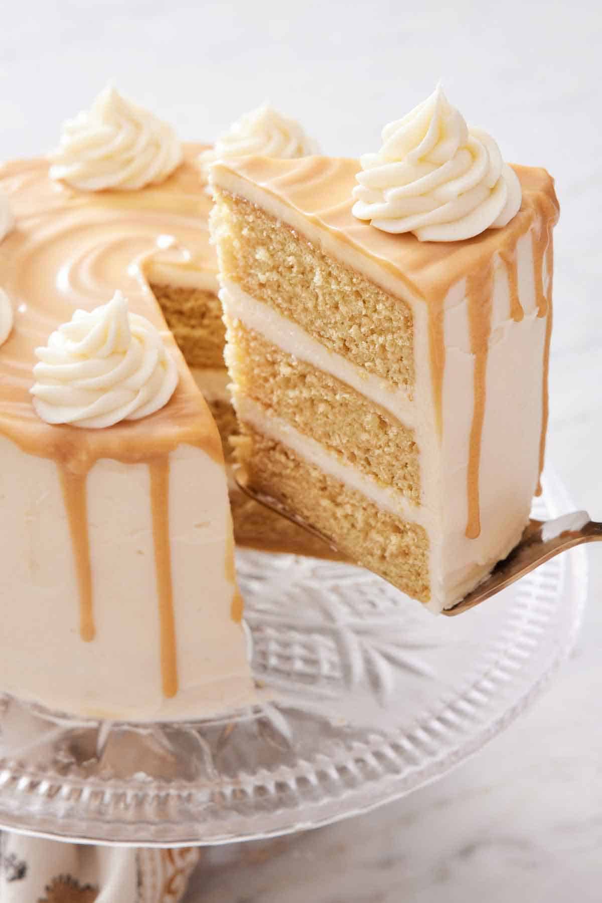 A slice of butterscotch cake being lifted from the cake on a cake stand with a spatula.