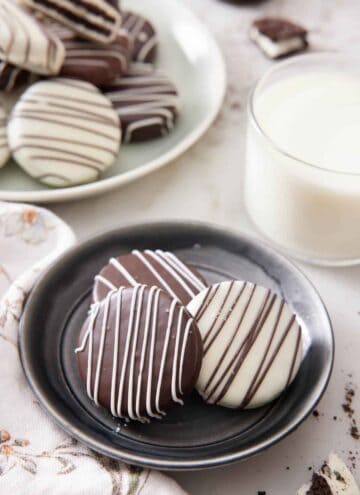 A plate with three chocolate covered oreos with a glass of milk and more oreos on a platter in the background.