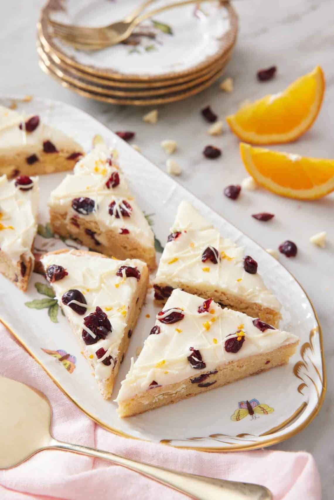 A platter with multiple pieces of cranberry bliss bars. A stack of plates in the background along with orange wedges.