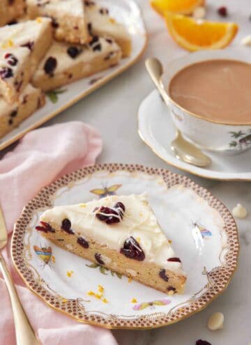 A plate with a slice of cranberry bliss bar with a coffee and platter with more bars in the background.