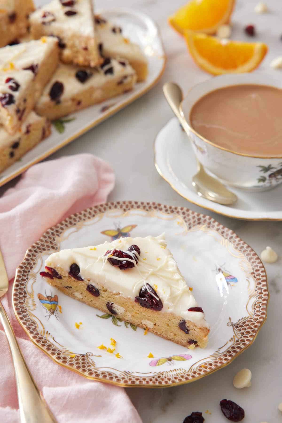 A plate with a slice of cranberry bliss bar with a coffee and platter with more bars in the background.