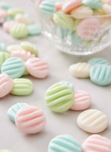 Multiple cream cheese mints in different colors on a marble surface with a bowl with more in the background.