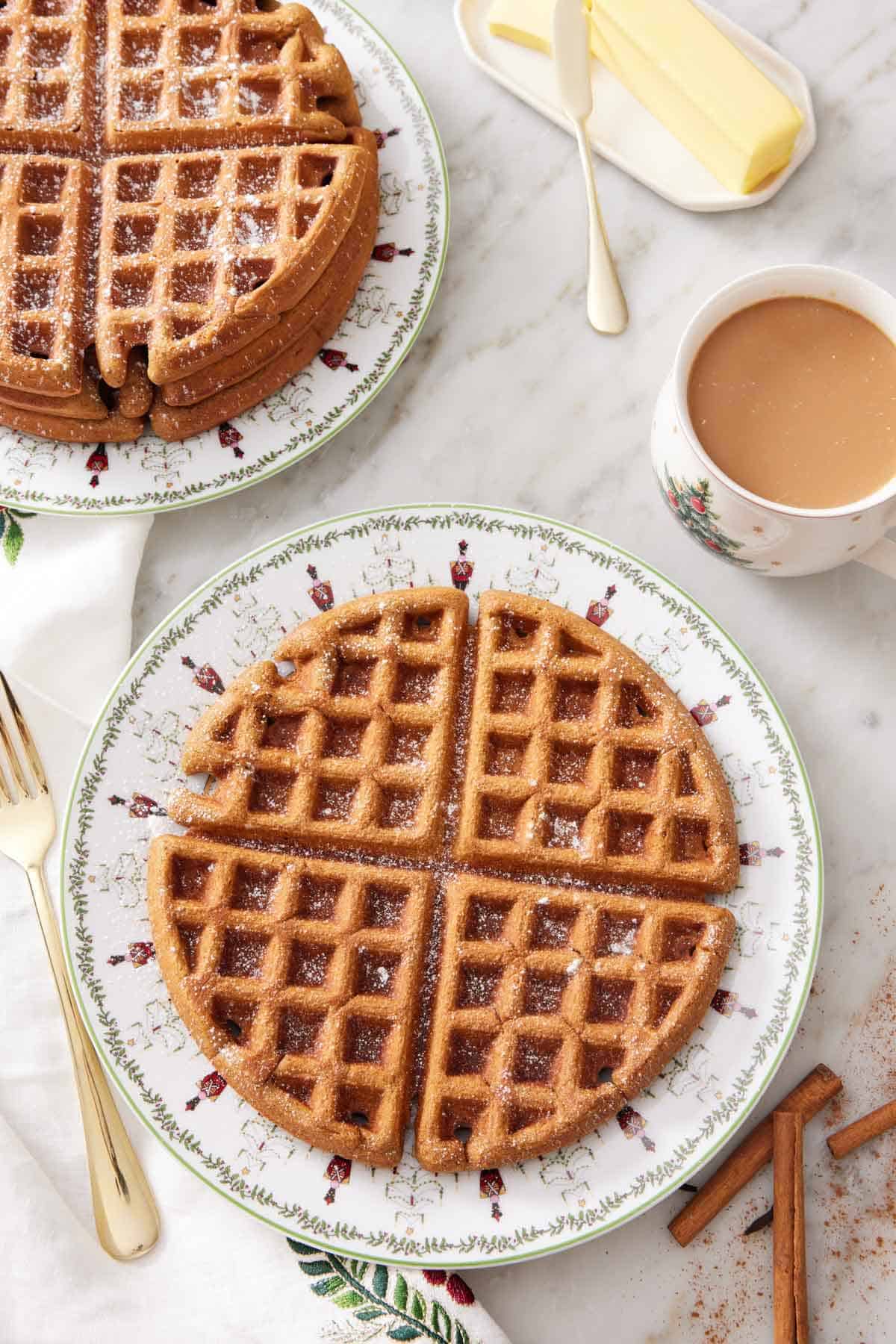 Overhead view of a large gingerbread waffle with a dusting of powdered sugar on top. A stack of waffles in the background along with a coffee and stick of butter.