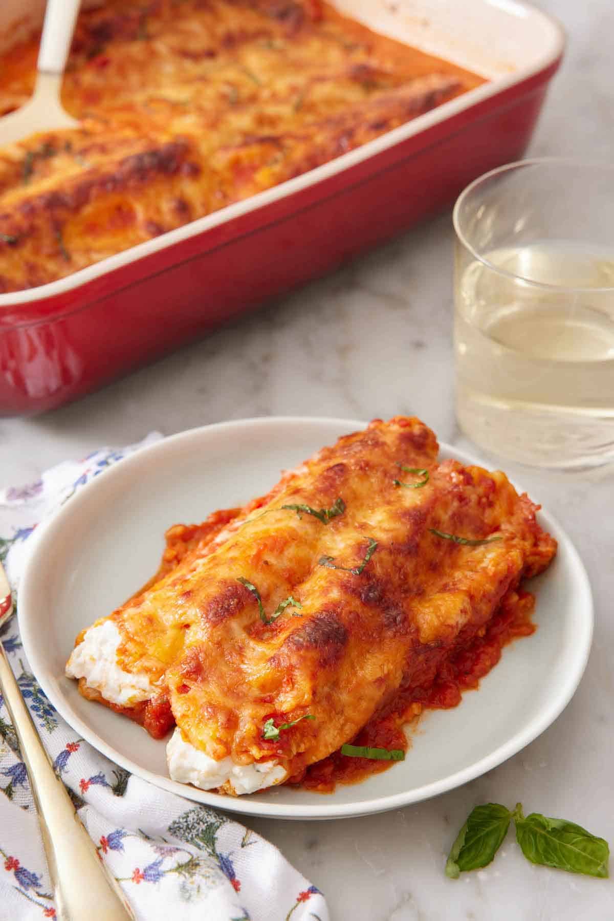 A plate with manicotti with a baking dish and glass of wine in the background.