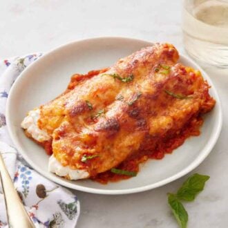 A plate with a serving of manicotti with a glass of wine and a fork beside it.