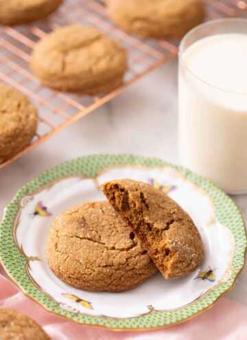 A plate with a molasses cookie with a half cookie slacked on top. A glass of milk in the background and more cookies on a cooling rack.