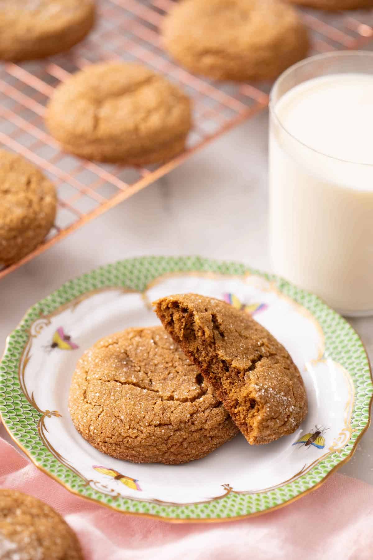 A plate with a molasses cookie with a half cookie slacked on top. A glass of milk in the background and more cookies on a cooling rack.