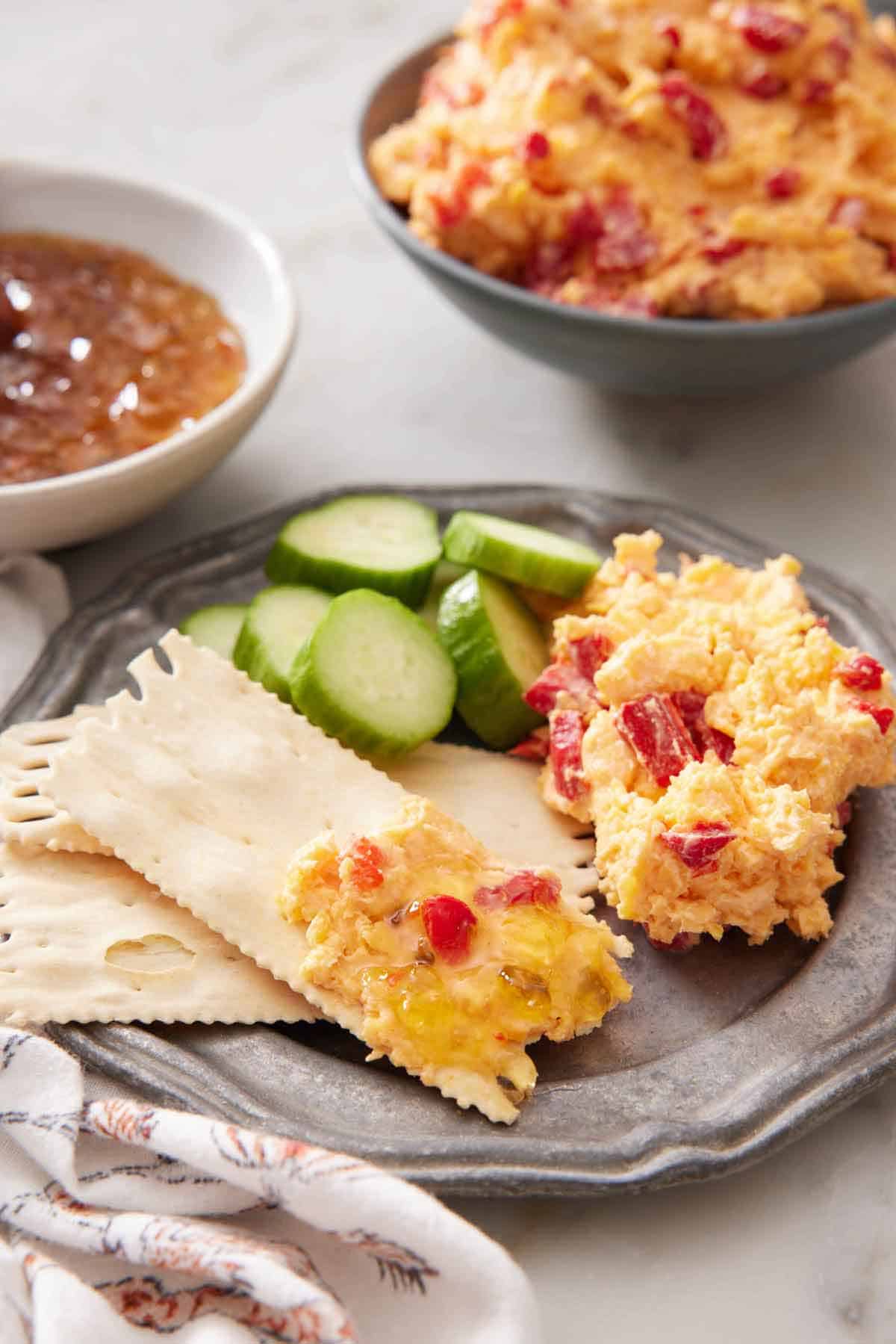 A plate pimento cheese with crackers with some cheese on one and sliced cucumbers beside them.