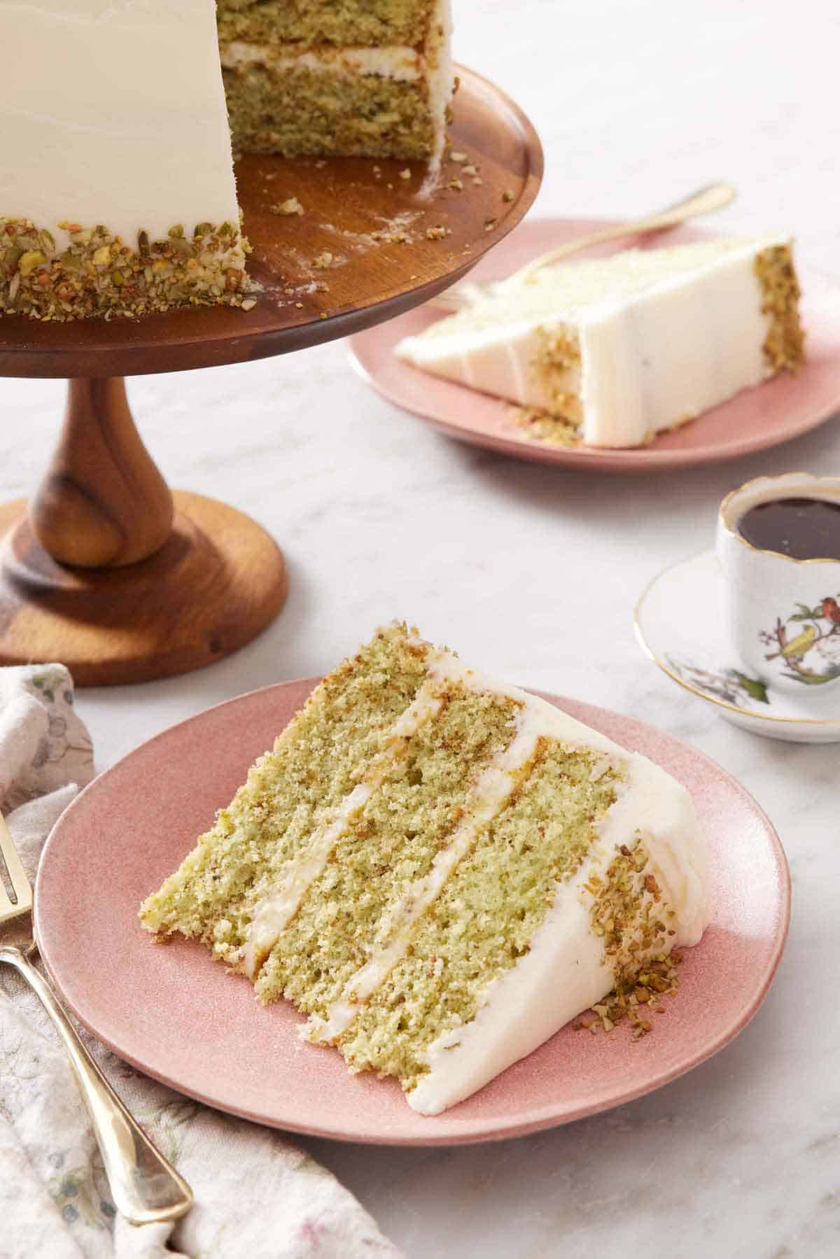 A slice of pistachio cake on a plate with the rest of the cake on a cake stand in the back along with a coffee and second plated slice.