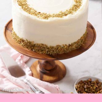 Pinterest graphic of a pistachio cake on a wooden cake stand with a bowl of shelled pistachios on the side.