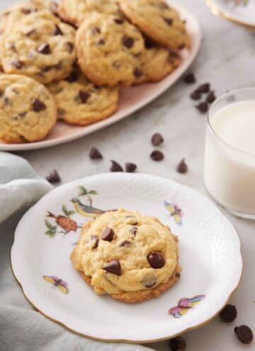 A plate with a pudding cookie with a platter in the background along with a cup of milk and chocolate chips scattered around.