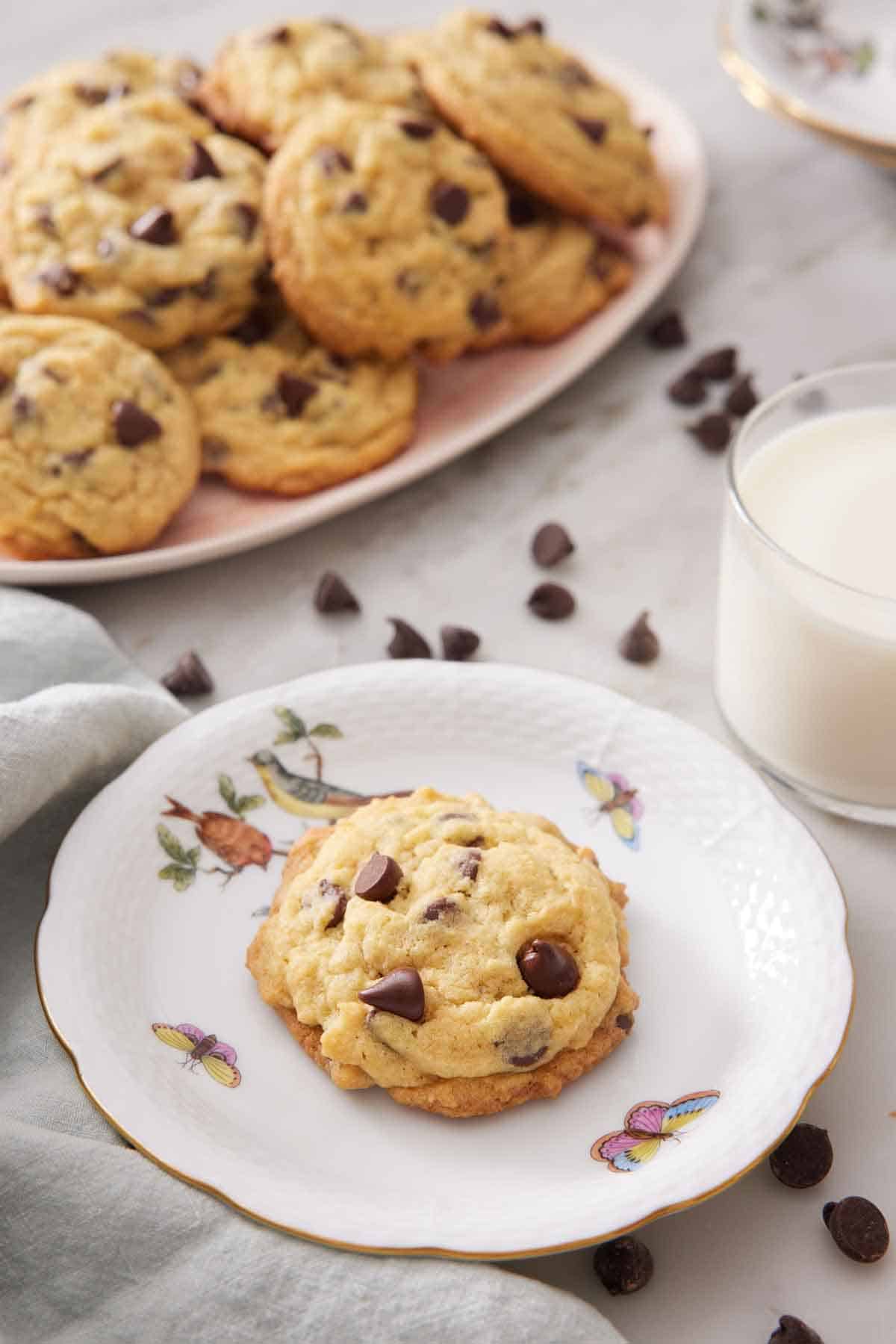 A plate with a pudding cookie with a platter in the background along with a cup of milk and chocolate chips scattered around.