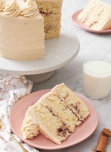 A slice of snickerdoodle cake with a glass of milk and the rest of the cake on a cake stand in the background.