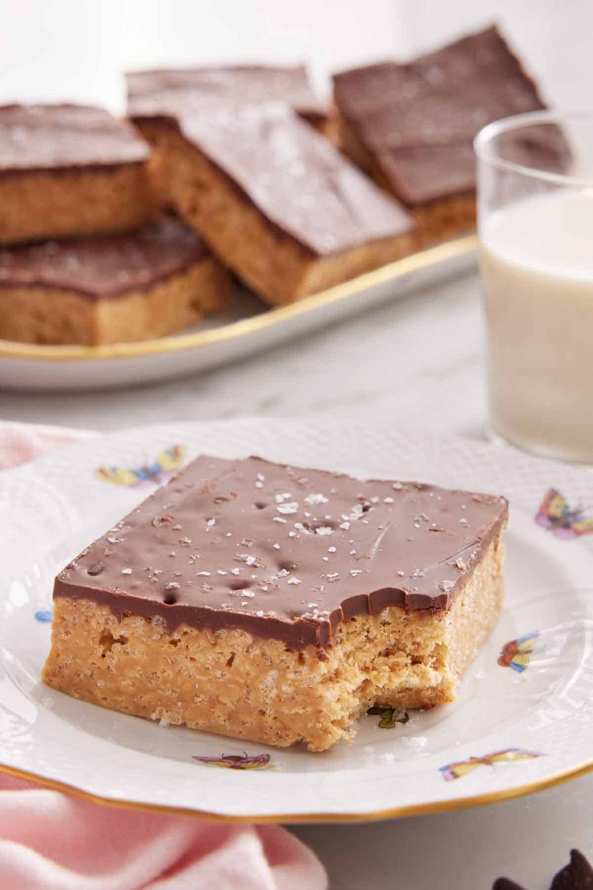 A plate with a Special K bar with a bite taken out of the corner. More bars in the background with a cup of milk.