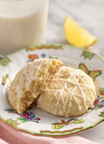 A plate with two lemon cookies, one cut in half. Lemon wedge and glass of milk in the background.