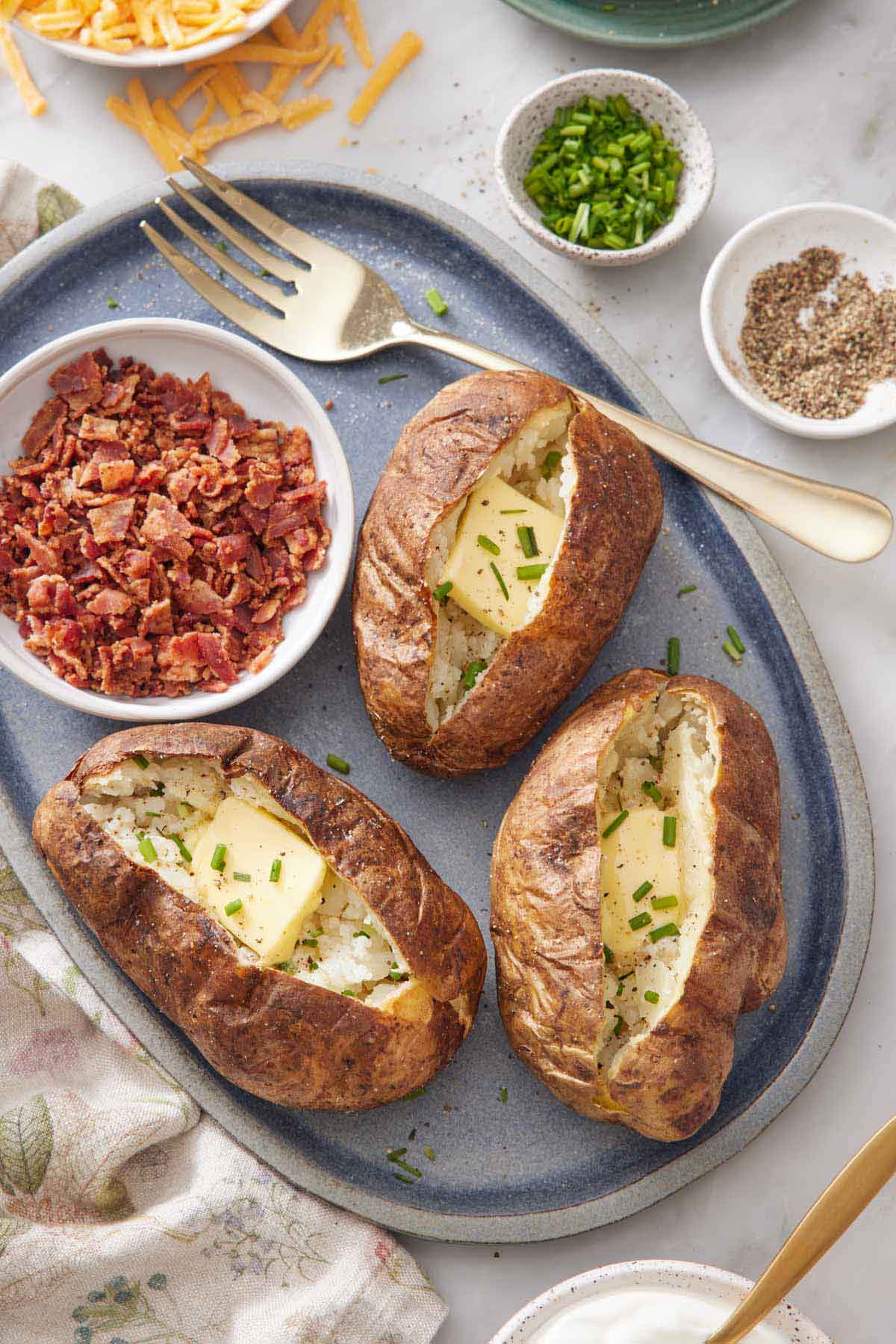 A platter with three air fryer baked potatoes topped with butter and chives with a bowl of crumbled bacon and a fork.