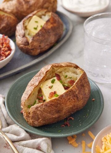 A plate with an air fryer baked potato topped with butter, chives, and crumbled bacon with more potatoes in the background.