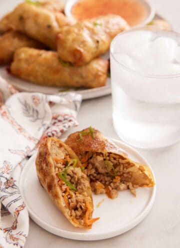 A plate with an air fryer egg roll cut in half. A glass of ice water and platter of more air fryer egg rolls in the background.