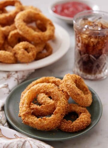 A plate of air fryer onion rings with a platter more in the back along with a glass of iced soda.