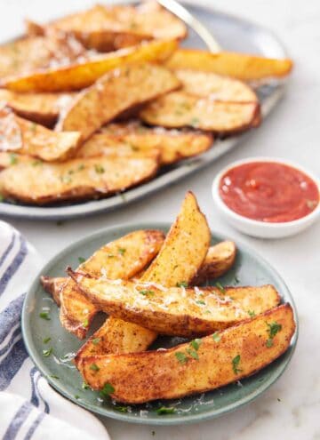 A plate of air fryer potato wedges topped with grated parmesan and parsley with a platter more in the background with a bowl of ketchup.
