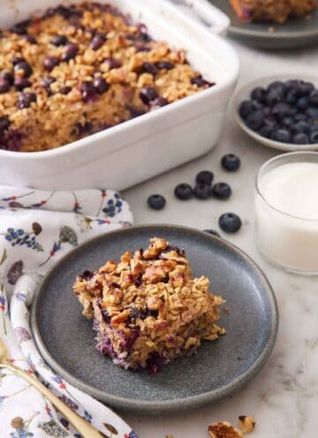 A plate with a square piece of baked oatmeal. Baking dish of the rest of the oatmeal, a bowl of blueberries, and a glass of milk in the background.
