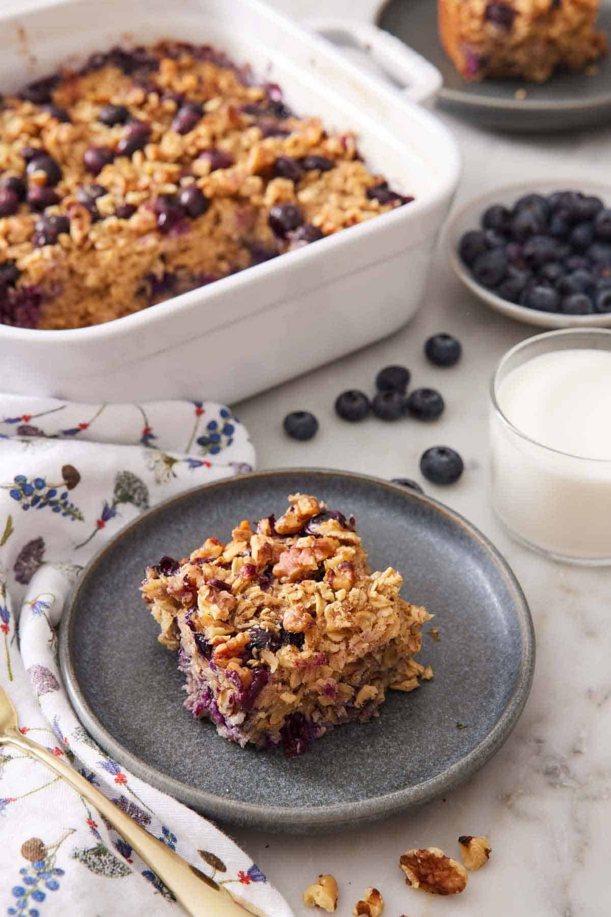 A plate with a square piece of baked oatmeal. Baking dish of the rest of the oatmeal, a bowl of blueberries, and a glass of milk in the background.