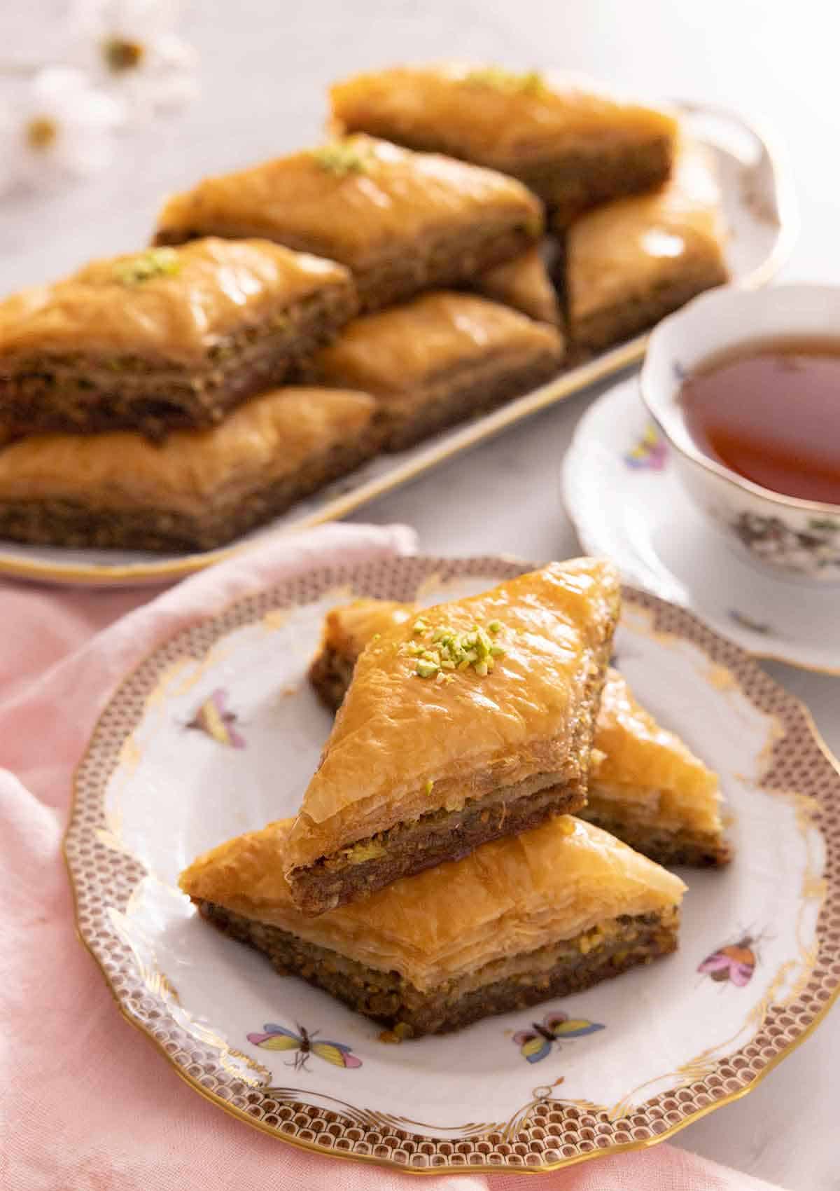 A plate with three pieces of baklava with a cup of tea and tray of more baklava in the background.