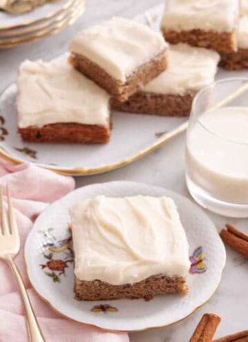 A plate with a slice of banana bar with a glass of milk and platter of more bars in the background.