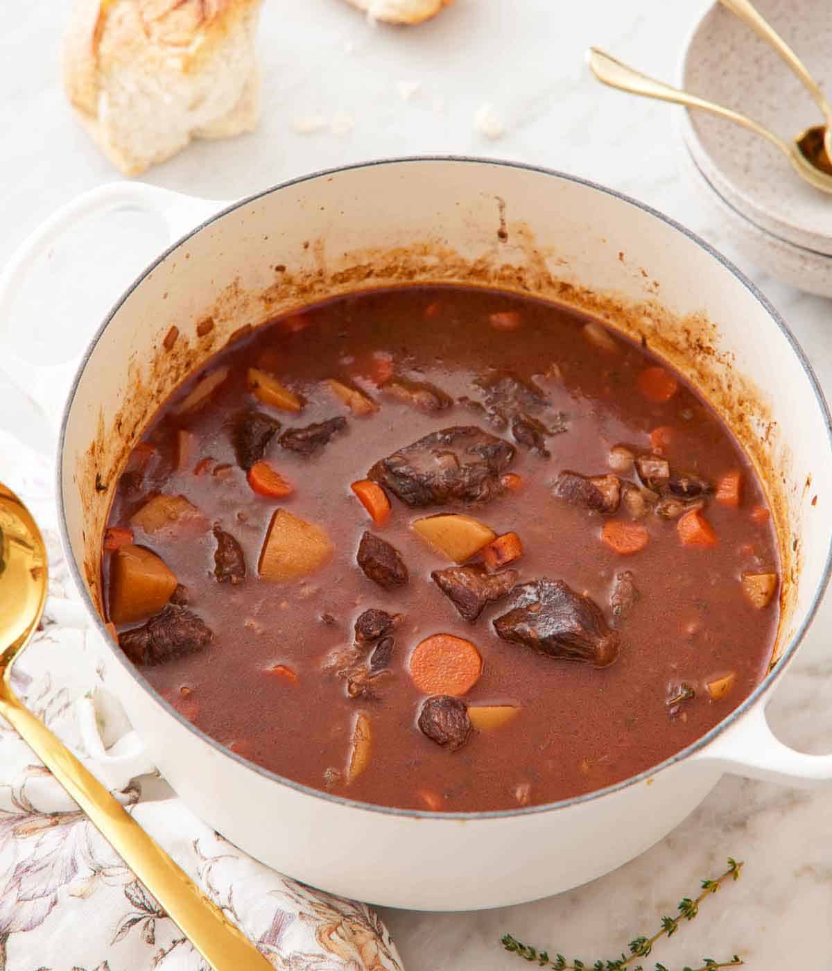 A white dutch oven with beef stew inside. Some torn bread and bowls in the background.