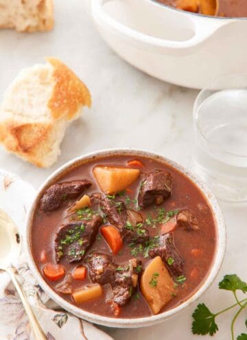 A bowl of beef stew with some torn bread, a glass of water, and a pot in the background.