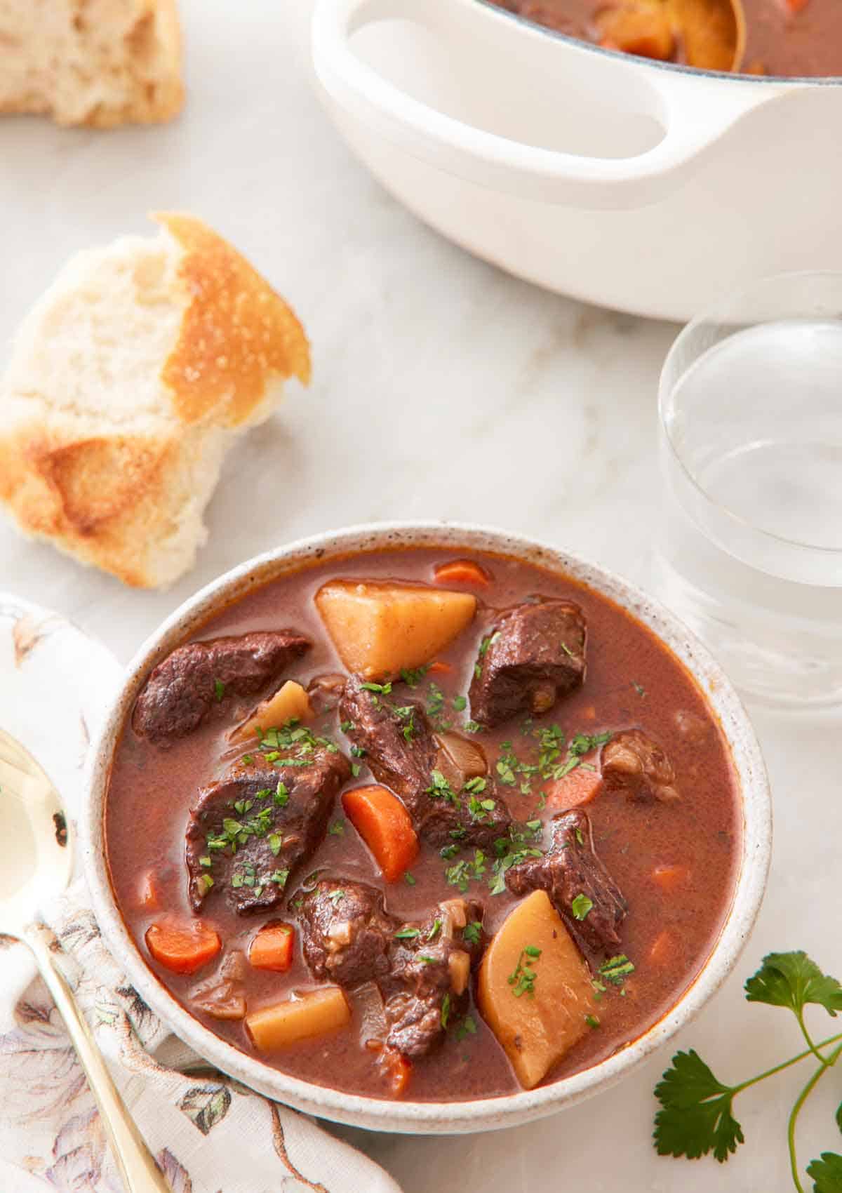 A bowl of beef stew with some torn bread, a glass of water, and a pot in the background.