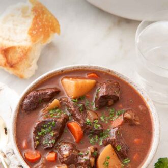 Pinterest graphic of a bowl of beef stew with bread, water, and a pot in the background.