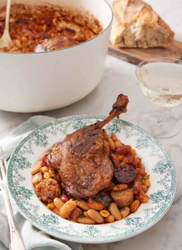 A plate with a serving of cassoulet with a pot with the rest in the background with some wine and bread.