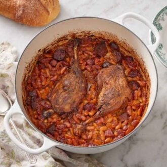 Overhead view of a white pot of cassoulet with a stack of bowls and forks on the side along with bread and a serving spoon on a linen napkin.