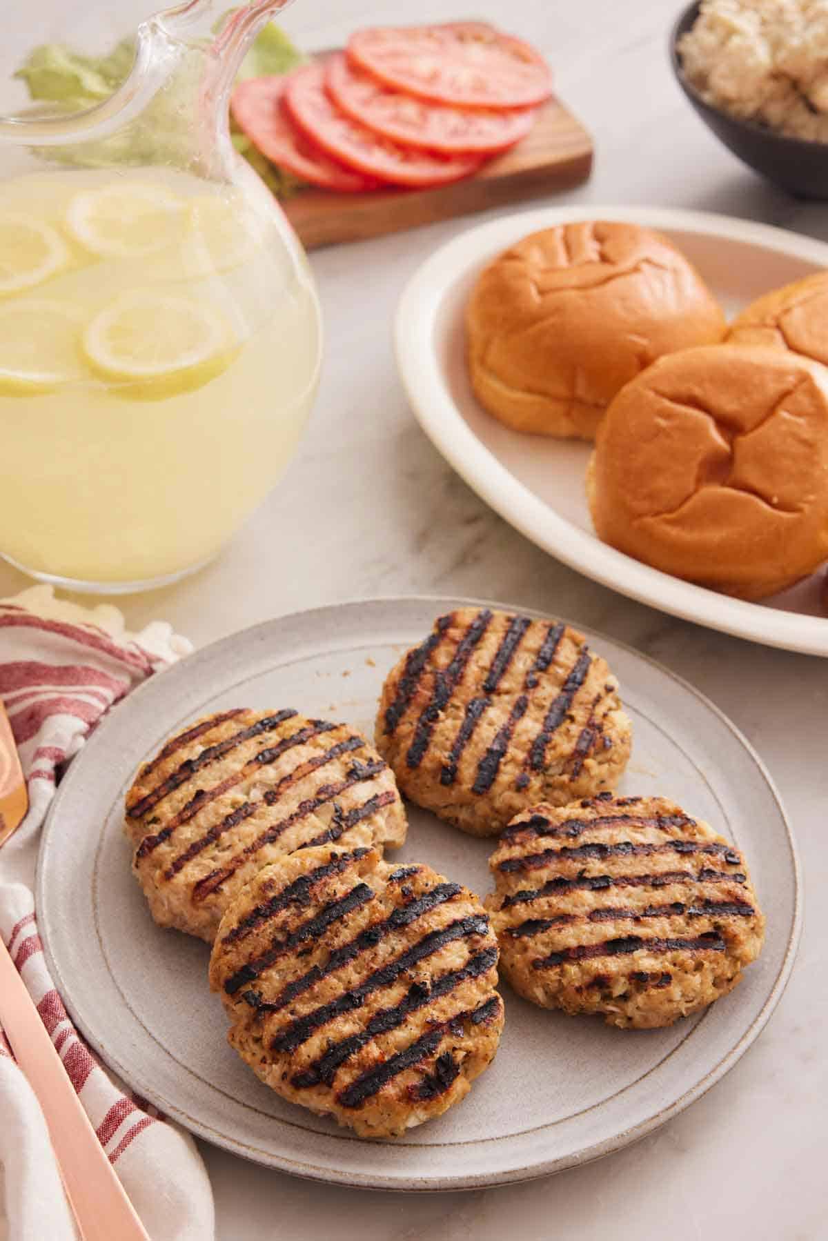 A plate with four chicken burger patties with a platter in the background with hamburger buns, a pitcher of lemonade, and more burger toppings.