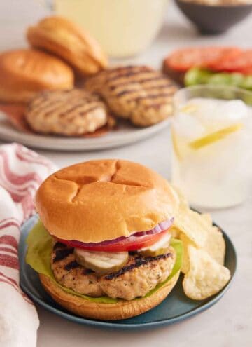 A plate with a chicken burger with potato chips with a drink in the background and burger components on plates.